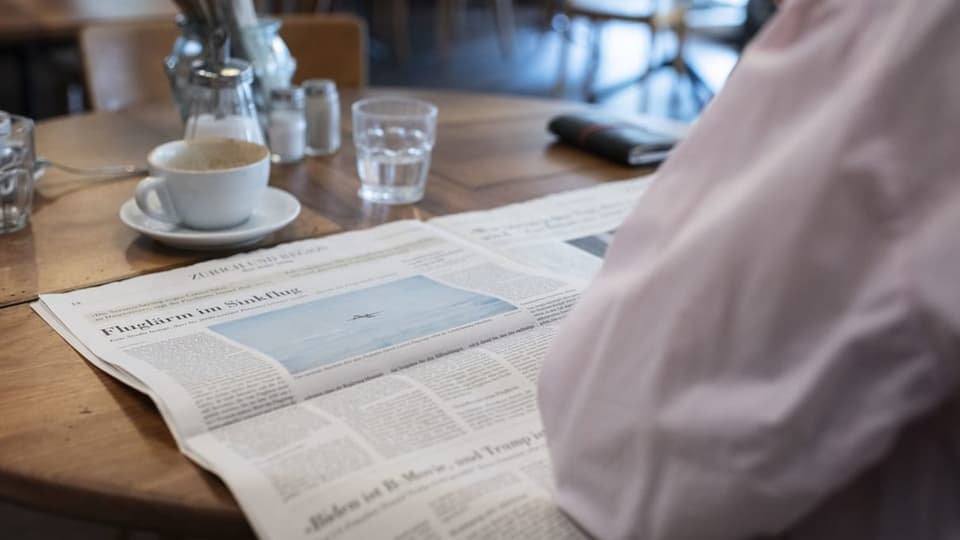 Zeitung liegt aufgeschlagen auf einem Holztisch in einem Restaurant, dahinter leere Kaffeetasse