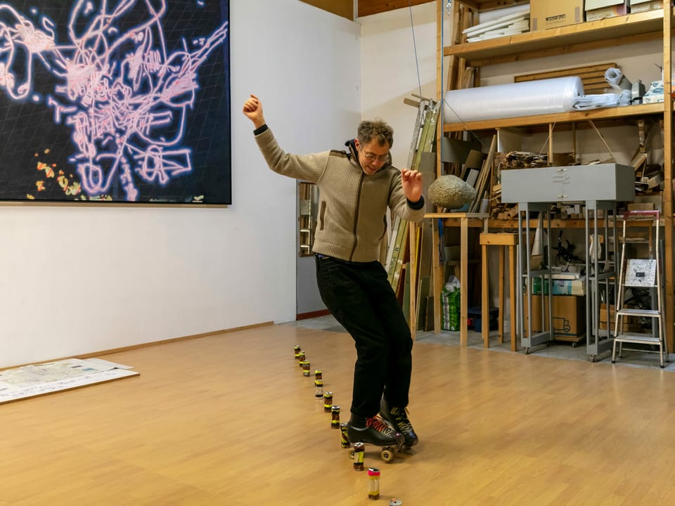 Ein Mann fährt in einem grossen Atelier-Raumauf Rollschuhen Slalom um in einer Reihe aufgestellte Dosen.