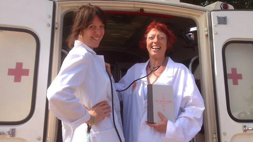Zwei Frauen in weissen Kitteln mit einem Buch stehen lachend vor einem Krankenwagen.