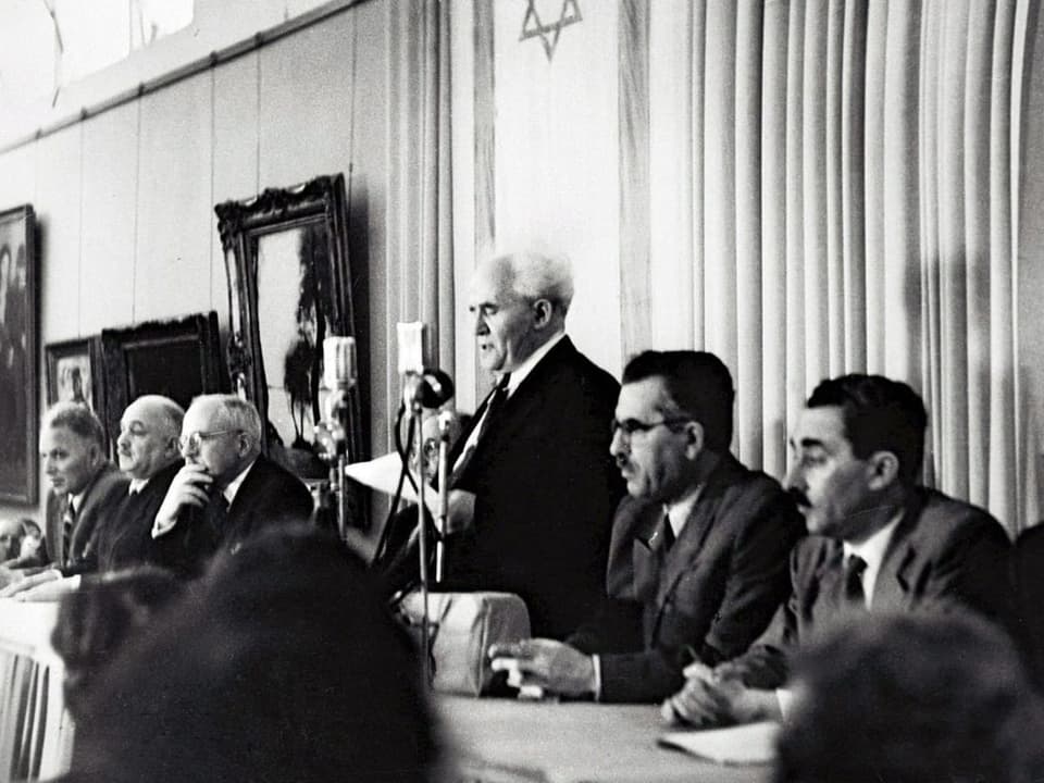 Ein schwarz/weiss Foto. Männer sitzen an einem Tisch. Gurion liest die Unabhängigkeitserklärung.