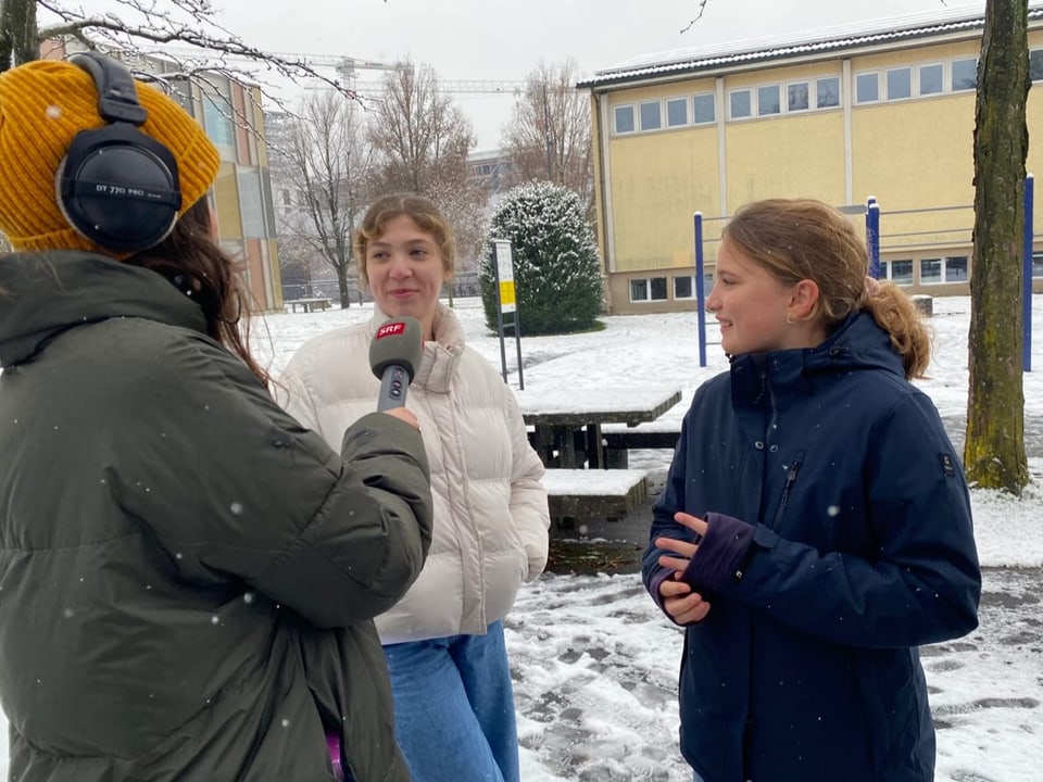 Zwei Mädchen antworten einer Frau mit Mikrofon auf dem Pausenplatz