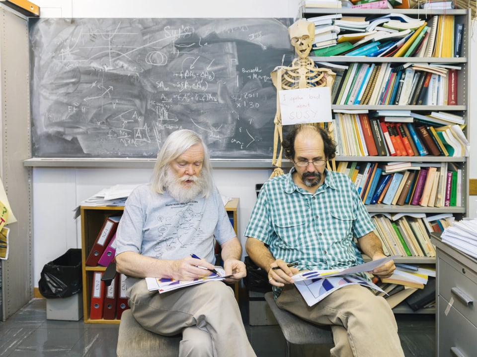 Aufnahme von zwei Forschern, die lesend vor einer vollgeschriebenen Tafel sitzen.