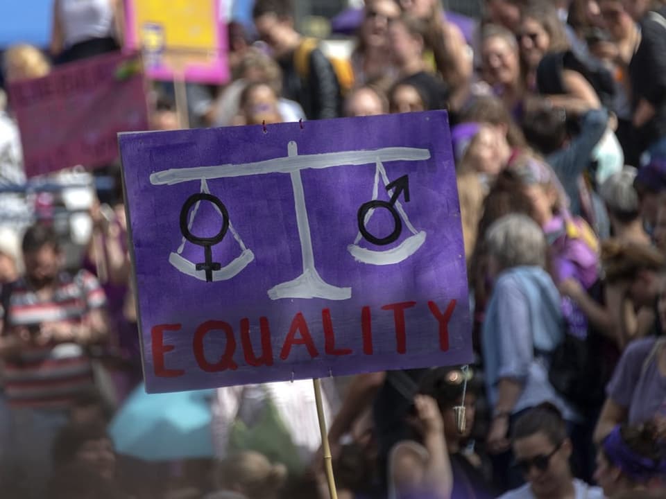 Demoplakat mit Waage, wo auf einer Seite ein Frauen- und auf der andern Seite ein Männerzeichen ist.