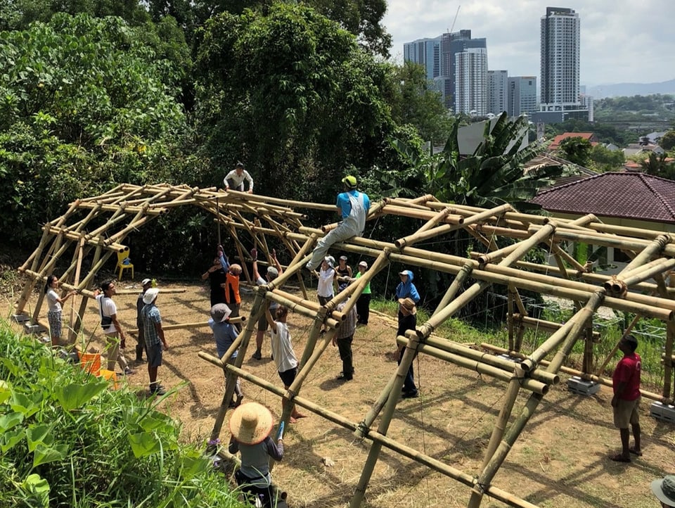 Dutzende Männer arbeiten auf einem Rohbau aus Bambus. Im Hintergrund sind Wolkenkratzer zu sehen.