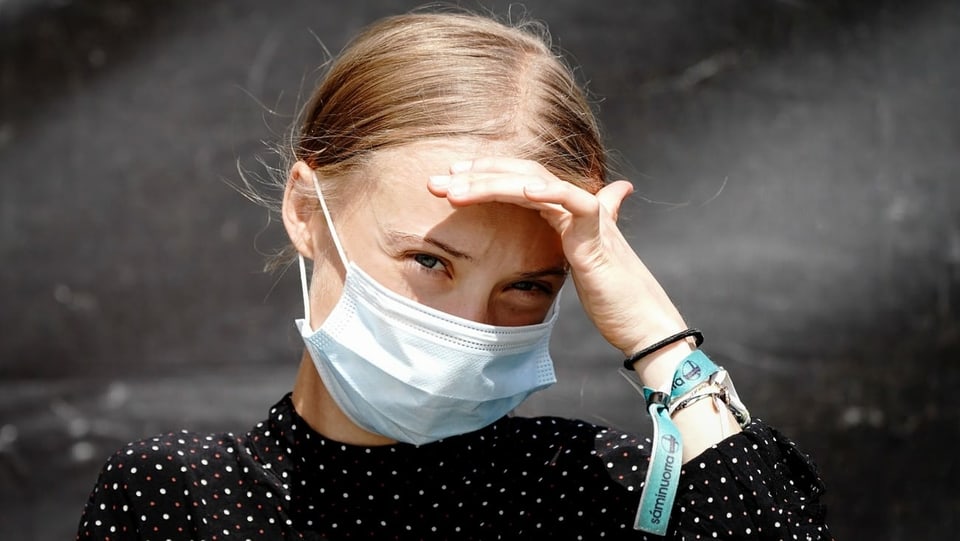Greta Thunberg trägt eine Hygienemaske und blickt ernst in die Kamera, eine Hand an der Stirn, um die Augen von der Sonne abzuschirmen.