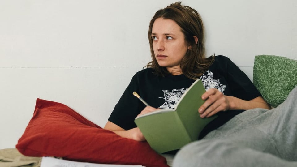Filmszene: Eine junge Frau in T-Shirt und Jogginghose sitzt auf dem Bett mit einem Notizbuch in der Hand.