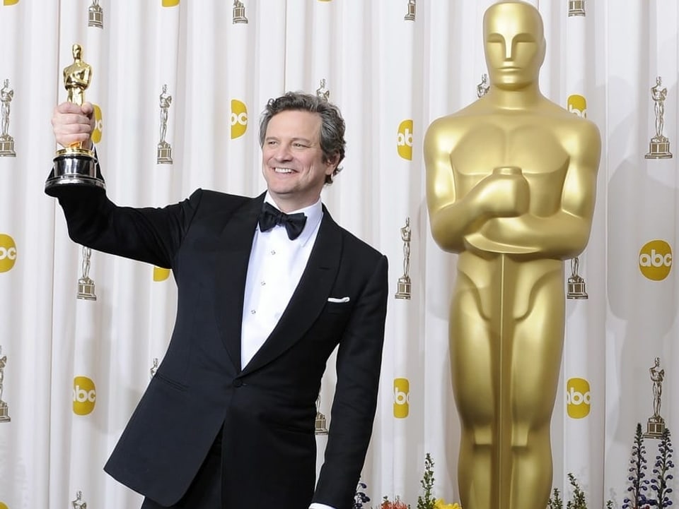 Colin Firth hält seinen Oscar in der rechten Hand und steht vor einer Oscar-Statue.