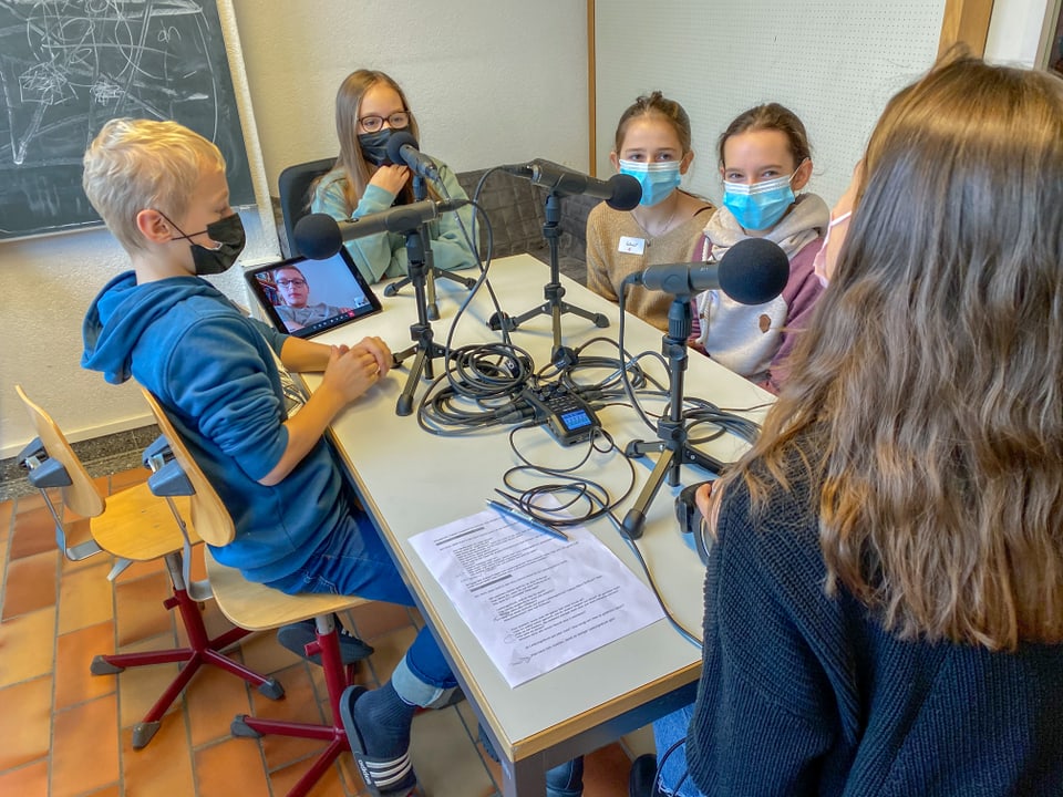 Kinder sitzen um einen Tisch, auf dem Mikrofone und iPad stehen. Darauf sieht man das Bild eines Jungen.