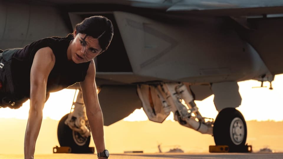 Filmszene: Eine Frau macht Liegestütze vor einem Kampfflugzeug.