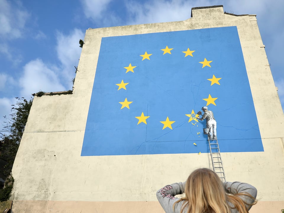 Darauf zu sehen ist eine mehrere Meter hohe quadratische EU-Flagge, aus der ein Handwerker einen Stern heraus meisselt. 
