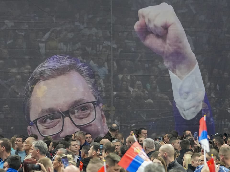 Menschenmenge teils mit serbischer Flagge, dahinter grosse Projektion von Mann mit Brille, reckt Faust in die Luft