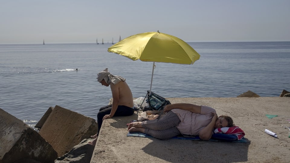 Ein Mann sitz unter einem gelben Sonnenschirm am Meer. Neben ihm liegt eine Frau im Schatten. 
