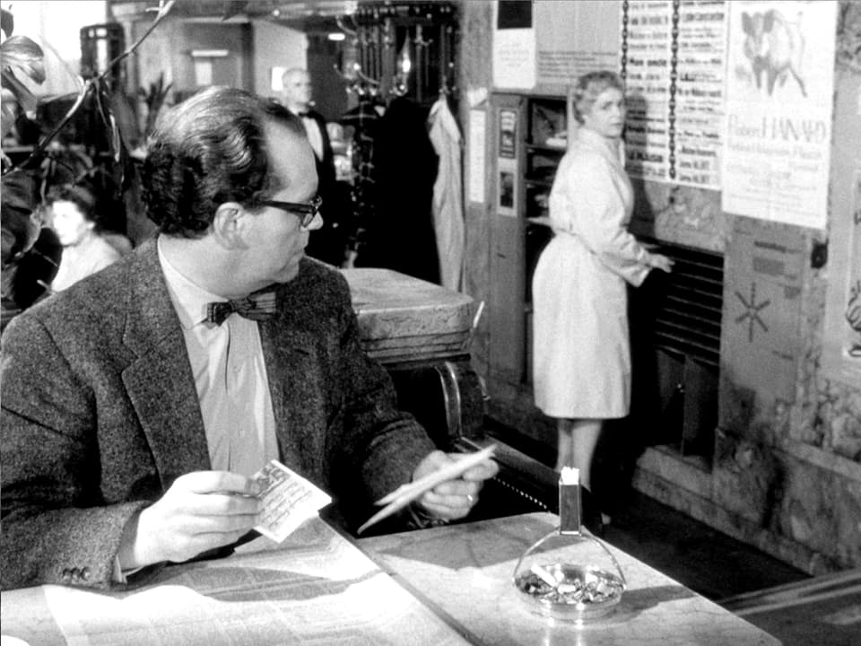 Ein Herr mir Fliege und Brille sitz an einem Tisch über seiner Zeitung. Er blickt jedoch zurück zu einer Frau, welche ein bisschen abseits von ihm an der Wand des Lokals steht.