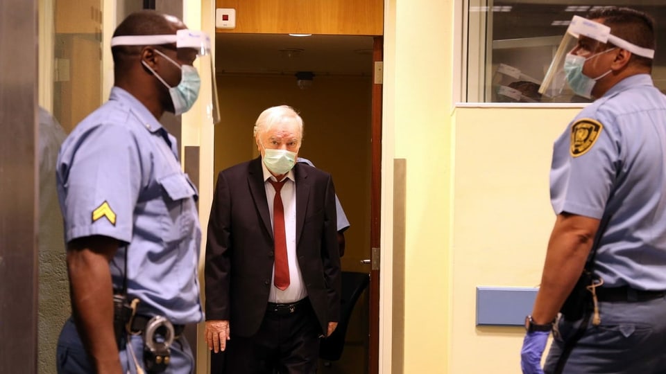 Mladic läuft in Anzug und mit Hygienemaske in den Gerichtssaal, links und rechts sind zwei Polizisten zu sehen.