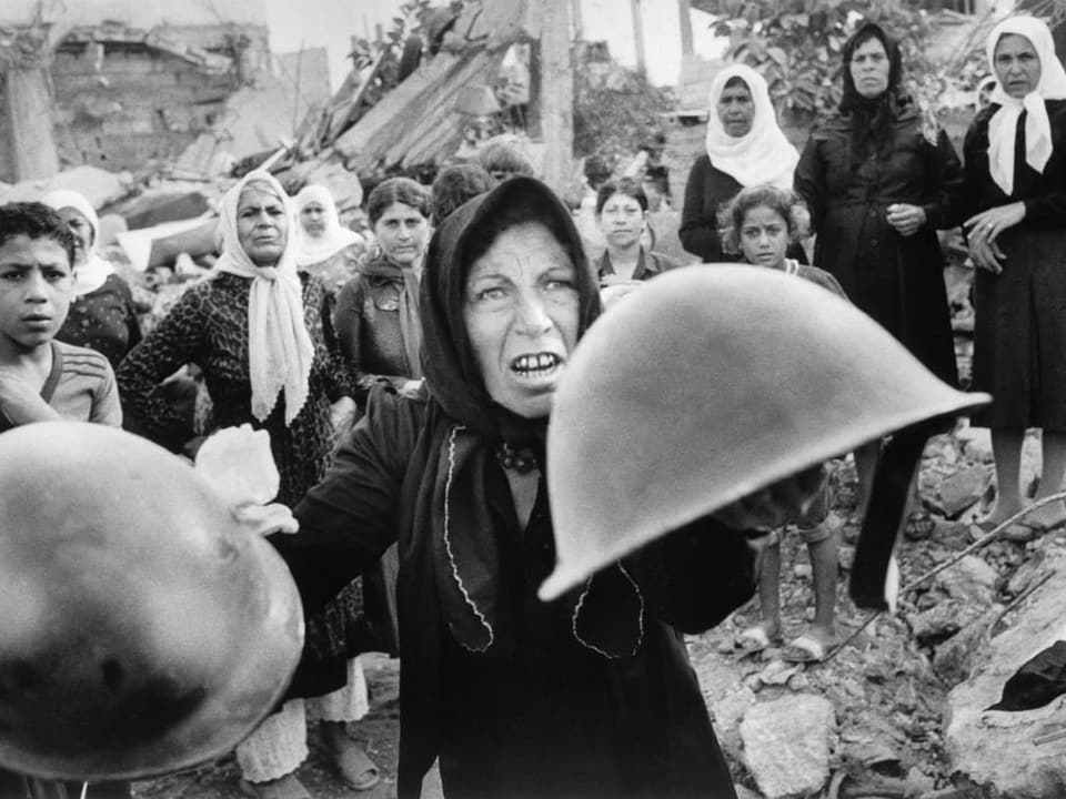Schwarz/weiss Foto: Eine Palästinenserin schwenkt Helme . Hinter ihr stehen weitere Personen in den Trümmern.