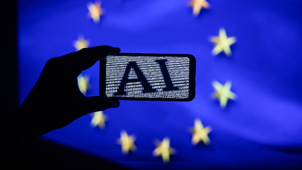 Eine Hand hält ein Handy mit einem AI-Bild in die Höhe, dahinter ist die EU-Flagge mit den goldenen Sternen zu sehen.