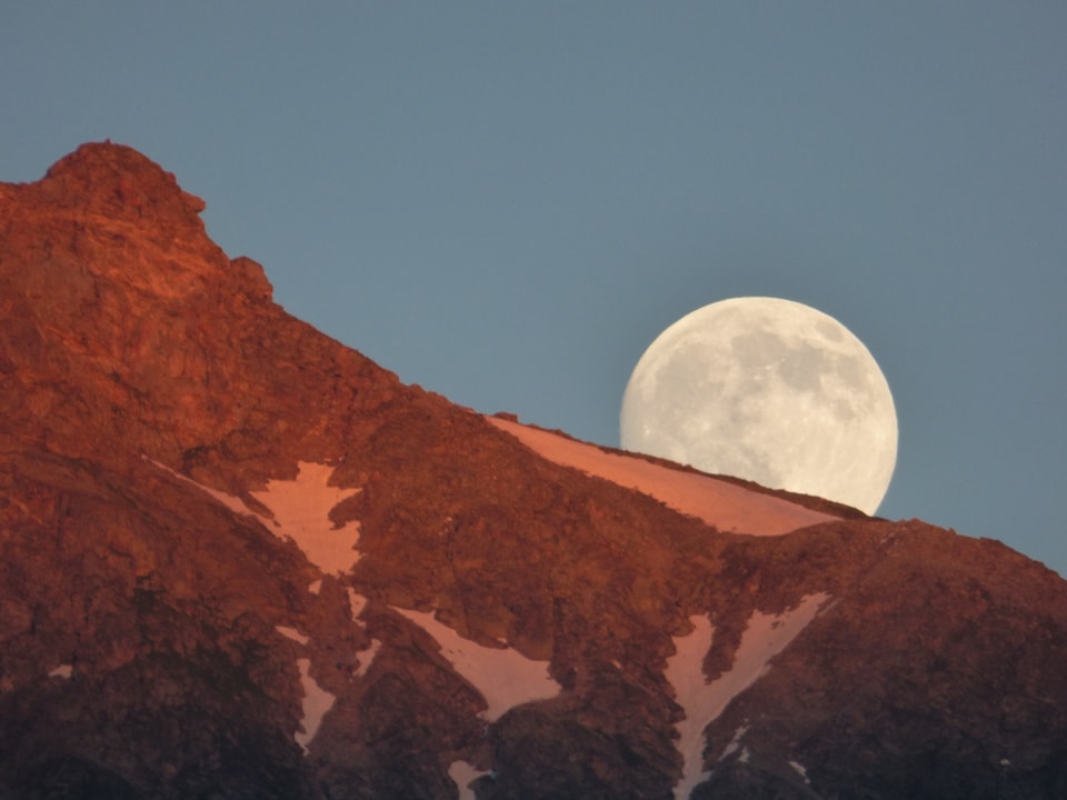 Der Vollmond geht über einer Bergflanke auf. Während die untergehende Sonne den Berg in eine rötliche Farbe taucht.