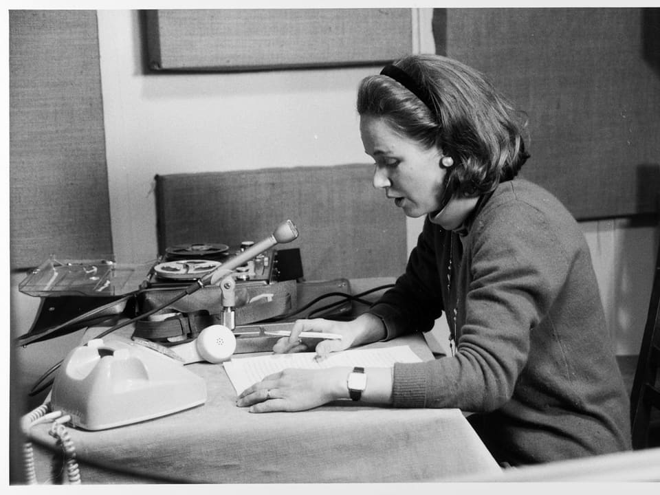 Frau sitzt vor einem Mikrofon, ein Telefonhörer liegt neben dem Apparat und im Hintergrund eine Bandmaschine.