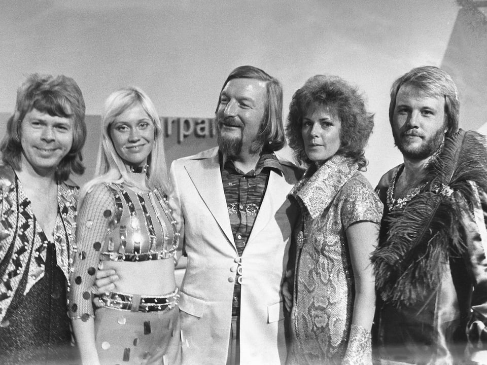 Schwarzweiss Bild mit James Last im weissen Sakko umrahmt von den ABBA-Gilrs und Jungs im wilden Siebziger Outfit. 
