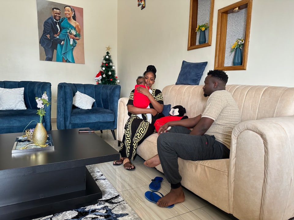 Blaise sitzt mit seinem Frau und dem Baby zuhause auf einem beigen Sofa. 