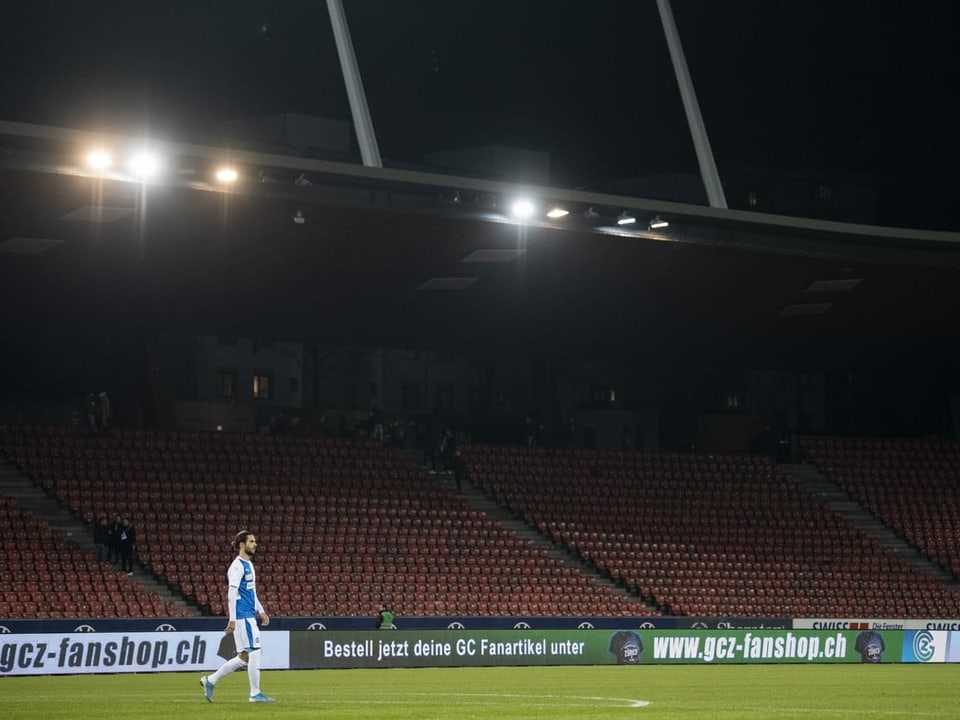 Ein Fussballer seht alleine auf dem grünen Rasen. Im Hintergrund die leeren Ränge eines Stadions. 