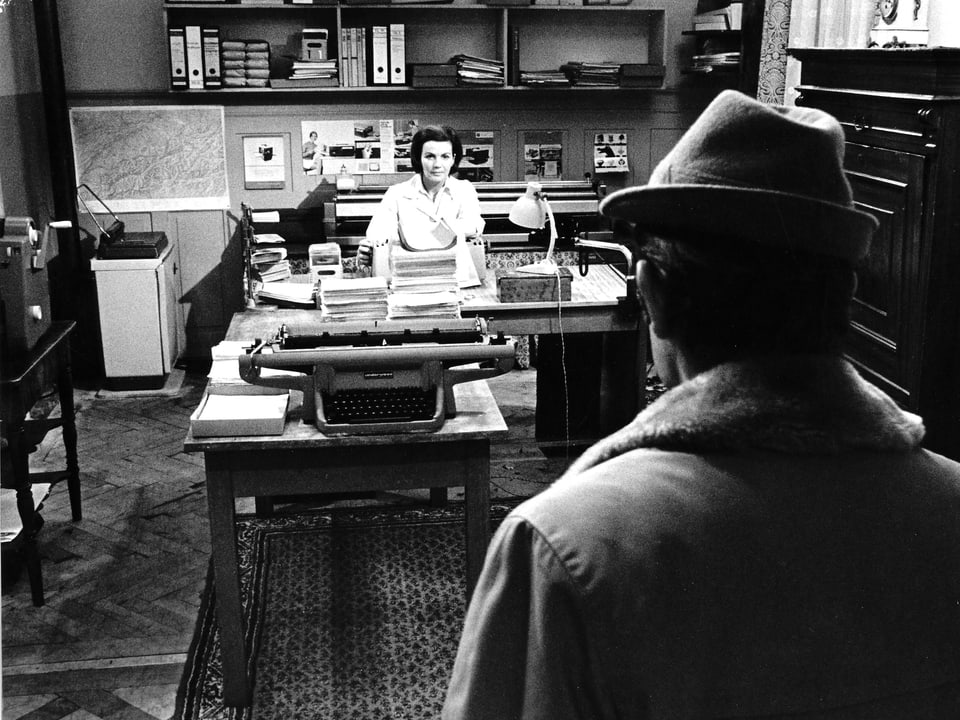 Eine Frau sitzt am Schreibtisch, davor steht ein Mann mit Hut.