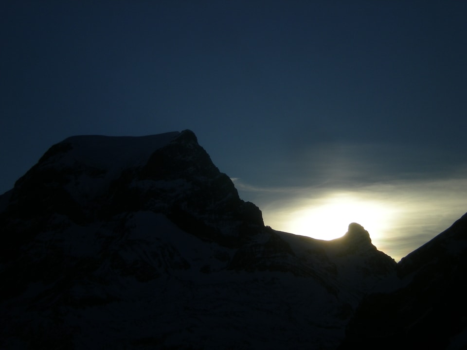 Der Himmel ist blau. Unten sieht man im Dunkeln ein Bergmassiv mit etwas Schnee. Die Sonne ist knapp hinter einer Bergkante zu erkennen.