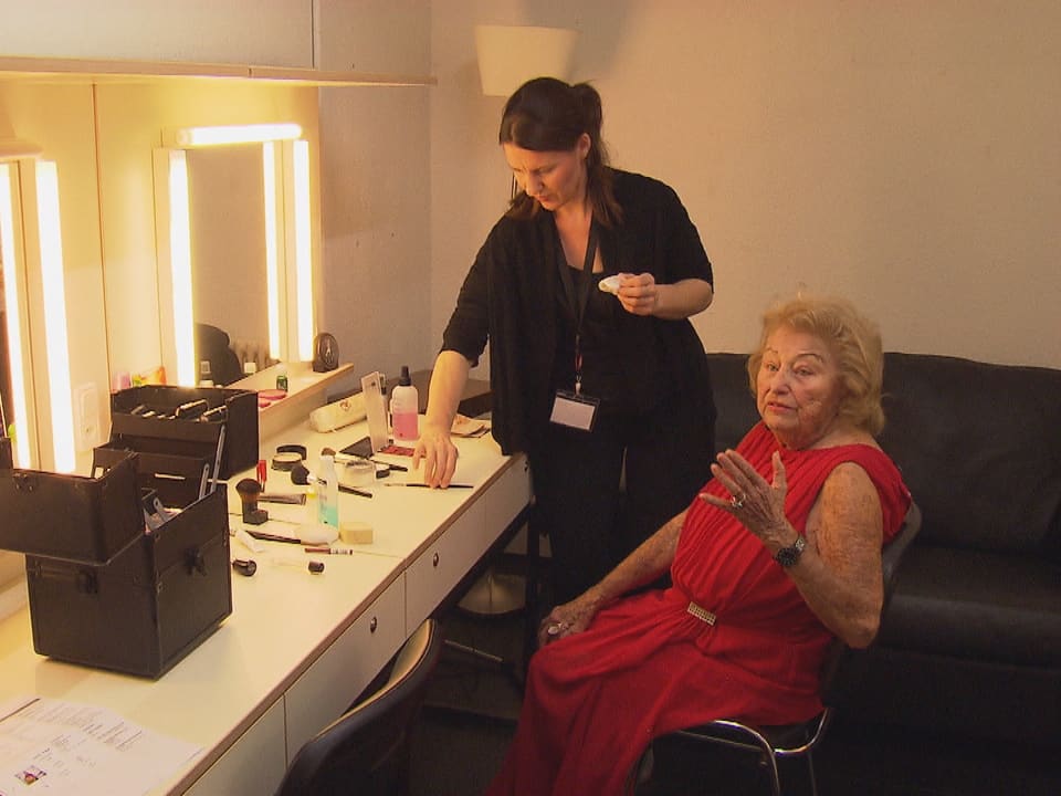 Eine ältere Frau sitzt vor einem grossen Spiegel und lässt sich schminken.