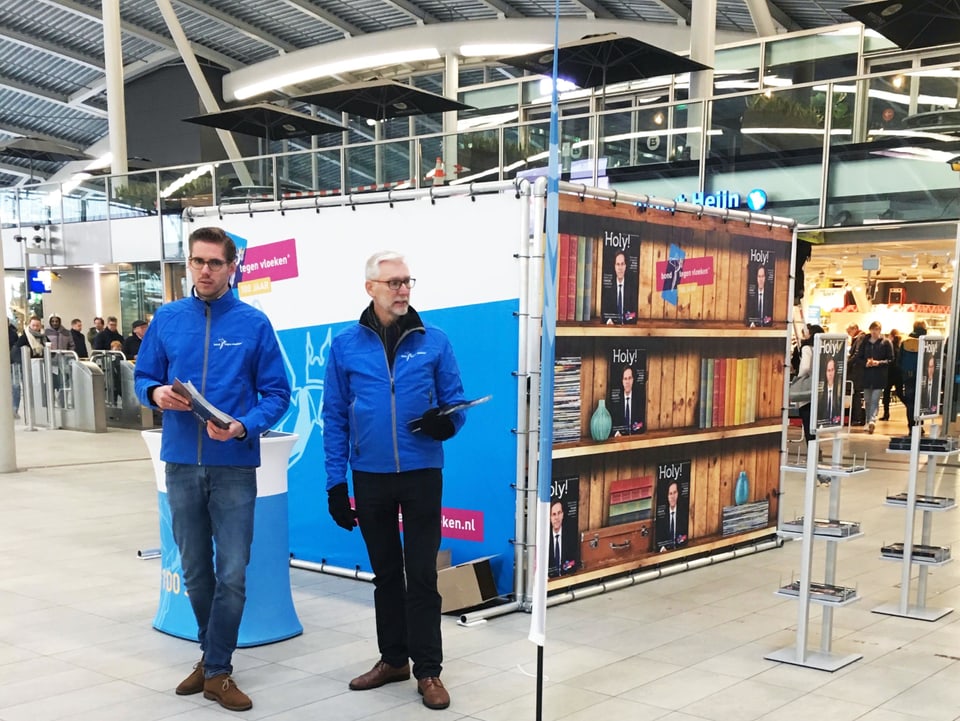 Zwei Männder in gleicher Jacke stehen an einem Stand in einer Halle.