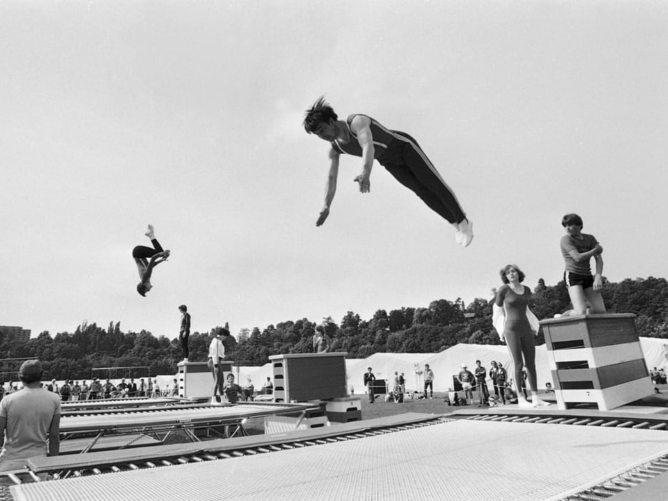 Das Foto ist schwarzweiss. Zu sehen sind zwei Kunstturn-Trampoline und zwei Männer in der Luft auf fast gleicher höhe. Einer befindet sich gerade mitten in einem Salto.
