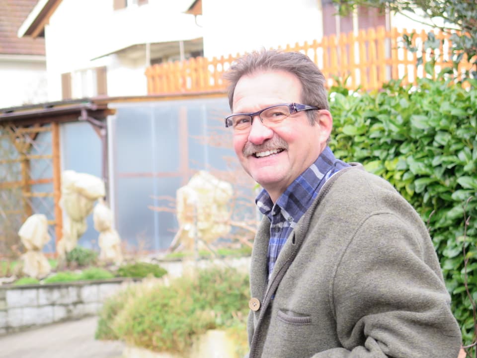 Mann in grauem Mantel lächelt, im Hintergrund ein Einfamilienhaus.