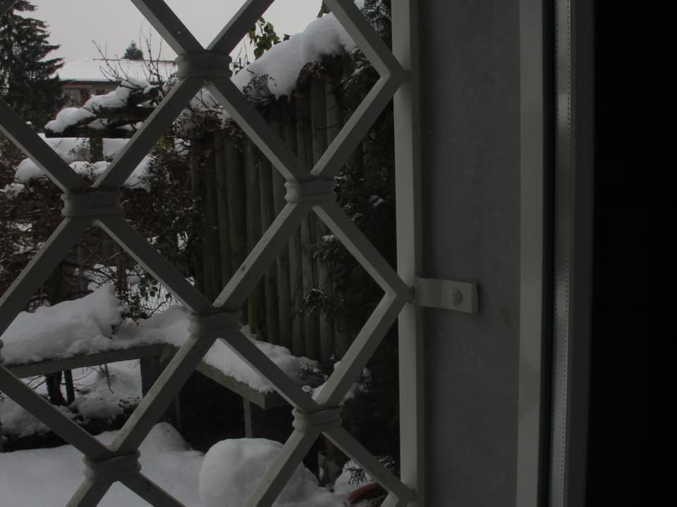 Blick aus einem vergitterten Fenster in den verschneiten Garten.