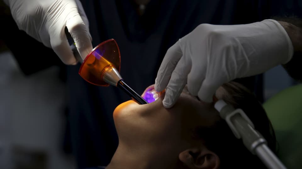 Sympbolbild: Patient auf dem Zahnarztstuhl, dieser hantiert mit Geräten im Mund des Patienten.