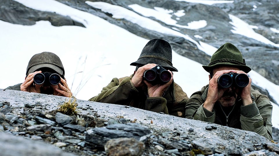 Drei Männer vin Jagdkleidung verstecken sich hinter einem Stein und schauen durch Ferngläser.