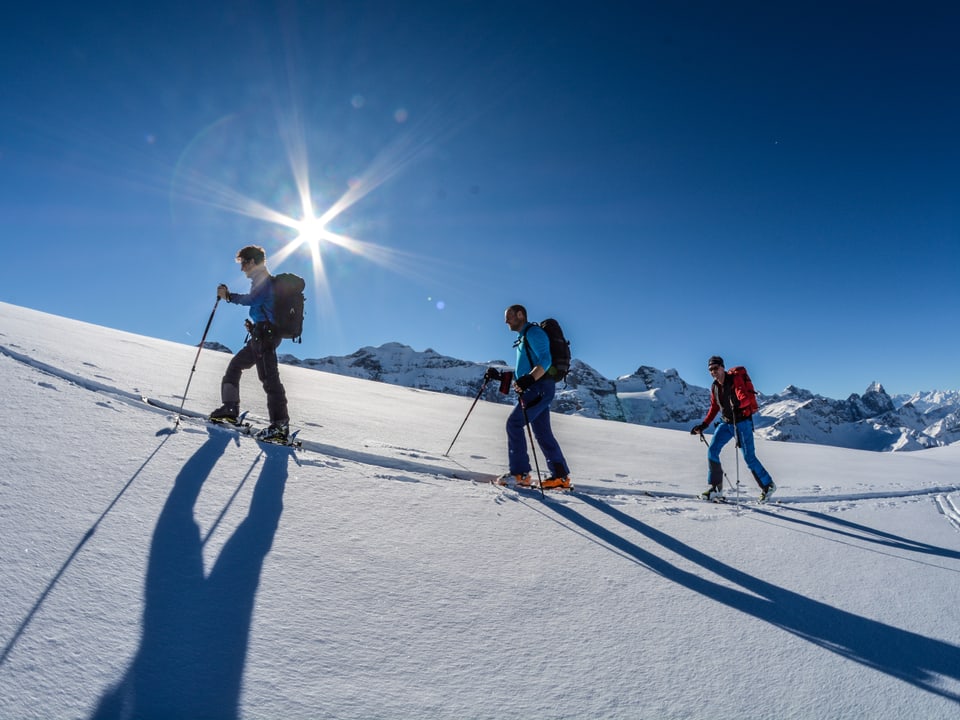 Drei Skitourenfahrer im Schnee, darüber blauer Himmel mit Sonne.