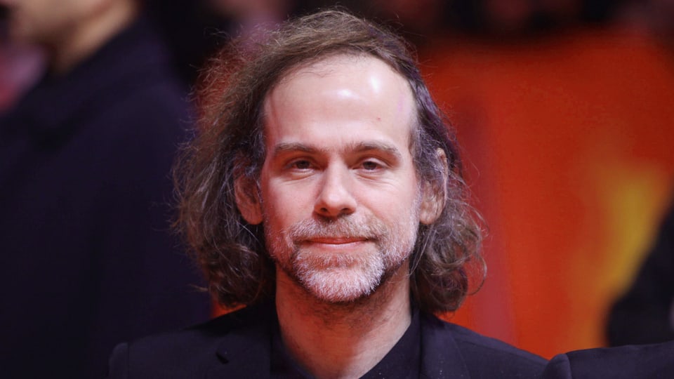 Mann mit längeren grauen Haaren (Porträt). Hintergrund rot.