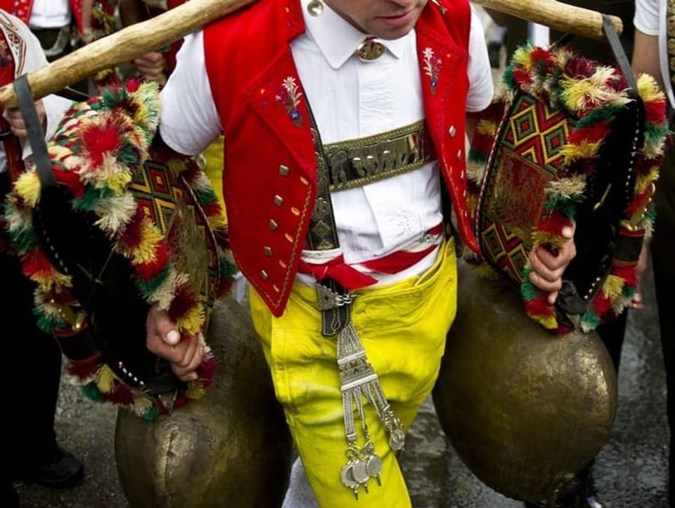 Ein Senn mit gelben Hosen, roter Weste und geschmückten Kuhglocken, die er auf den Schultern trägt.
