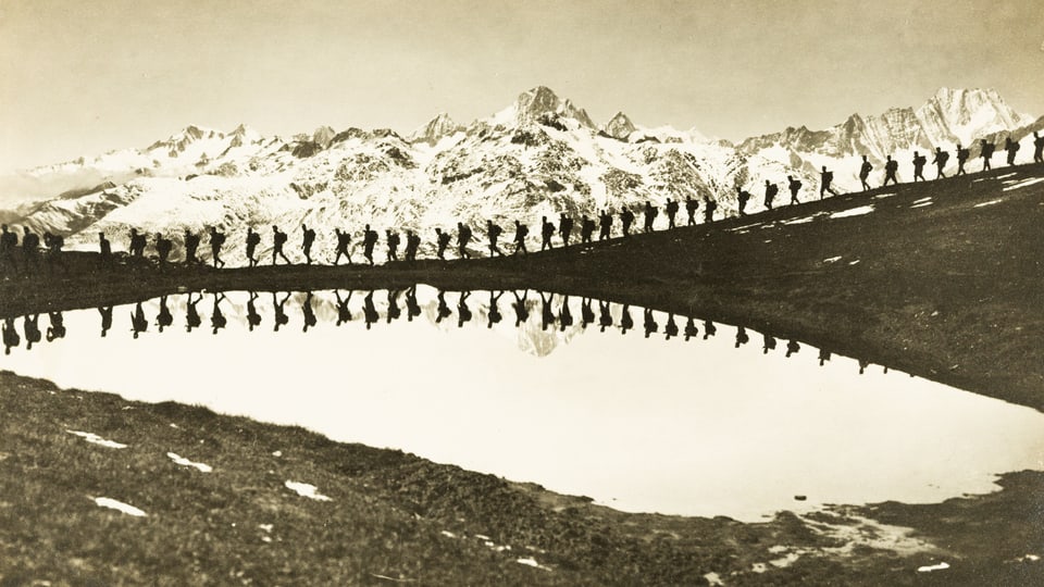 Viele Männer in einer Reihe spazieren an einem See entlang. Hinten sieht man Berge.