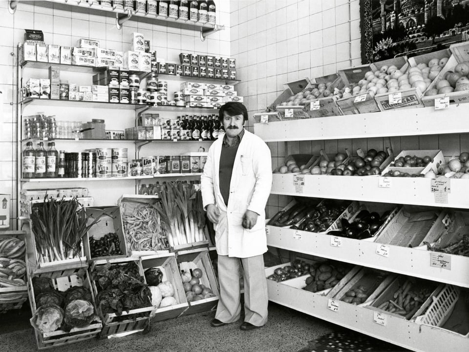 Ein türkischer Verkäufer in weissem Kittel steht vor Gemüse- und Früchteregalen.