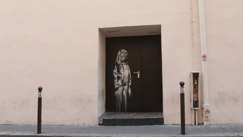 Cremefarbene Wand, darin eine schwarze Tür. Auf die Tür gesprayt, in Weiss: ein trauriges Mädchen, madonnenähnlich.