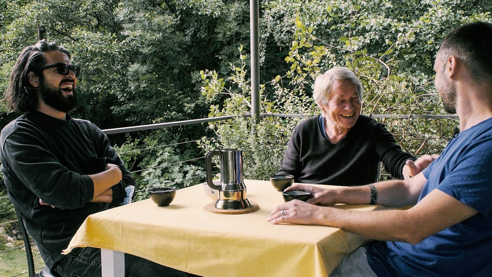Drei Männer lachend draussen an einem Tisch, auf dem eine Kaffekanne steht.