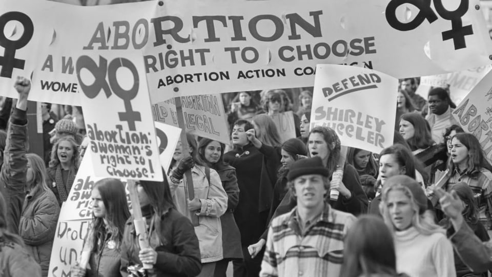 Schwarz-weiss Aufnahme eines Demonstrationszuges mit Frauen und Männern von 1970. Forderung für «Recht auf Abreibung».