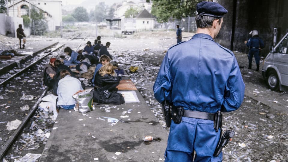 Polizist steht vor einer Gruppe Menschen, die auf völlig verdrecktem Untergrund sitzen.