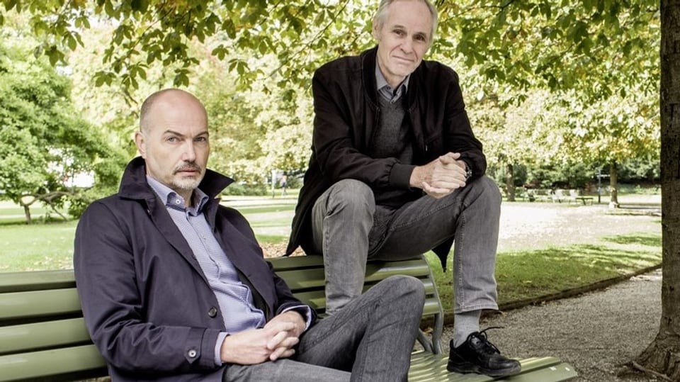 Zwei Männer sitzen auf einer Parkbank. Links ein Mann mit Halbglatze, rechts grauhaariger Mann.