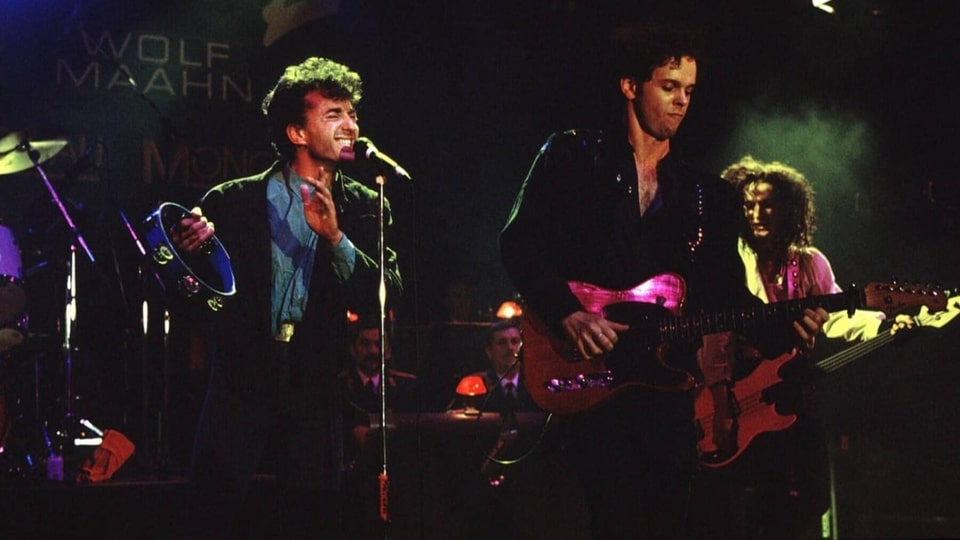 Farbfoto links in blauem Hemd singender Mann mit Locken, Mitte Mann mit Gitarre, rechts hinten Mann mit langen Locken