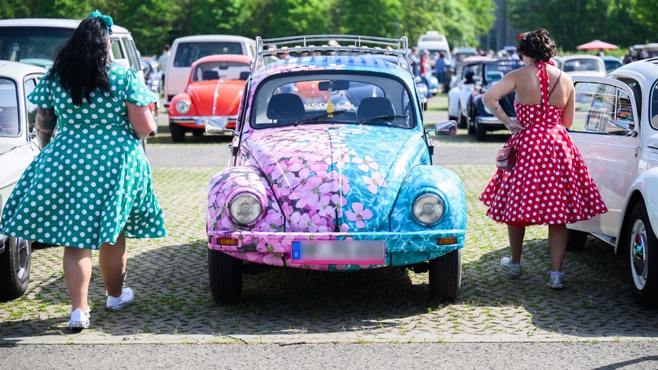 Zwei Frauen in gepunkteten Kleidern betrachten bunte Volkswagen Käfer auf einem Oldtimertreffen.