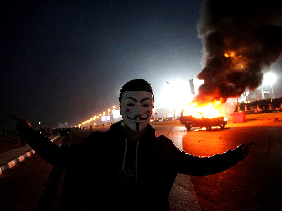 Fan mit Guy Fawkes Maske steht vor brennendem Auto