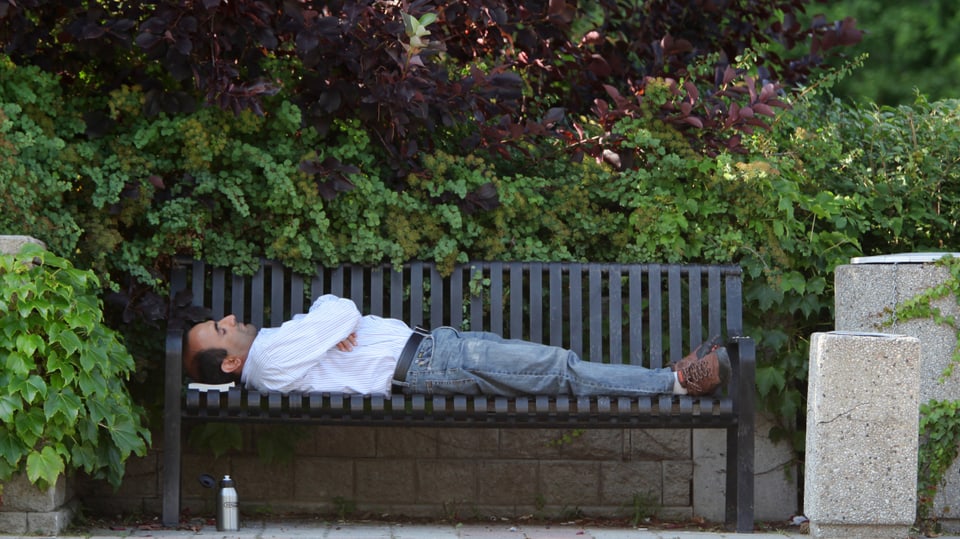 Ein Mann mit weissem Hemd und Jeans liegt auf einer Parkbank, die Arme verschränkt. Dahinter: Grüne und rötliche Büsche.