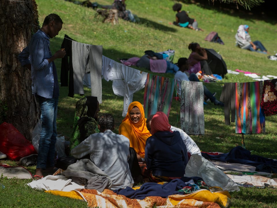 Flüchtlinge sitzen auf Decken in einem Park, dahinter Wäscheleinen