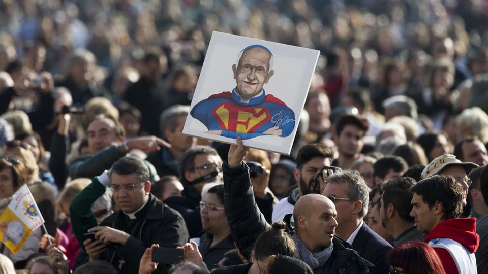 Eine Menschenmenge, aus der ein Mann heraussticht, der ein Plakat in die Luft hält, das den Papst als Superman zeigt.